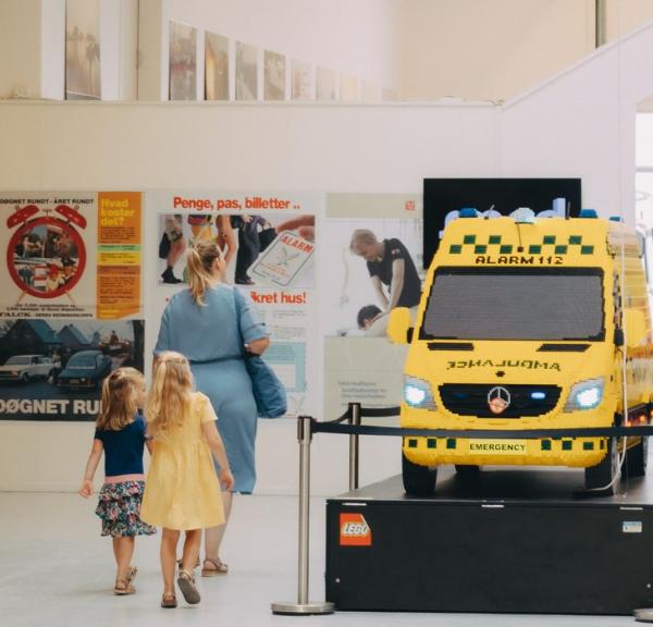 Ambulance og brandbil på indendørs udstilling. En mor med to børn går rundt og kigger på udstillingen.