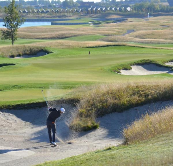 Golfspiller på bølgende grøn golfbane slår et slag fra en bunker, så sandet står op.