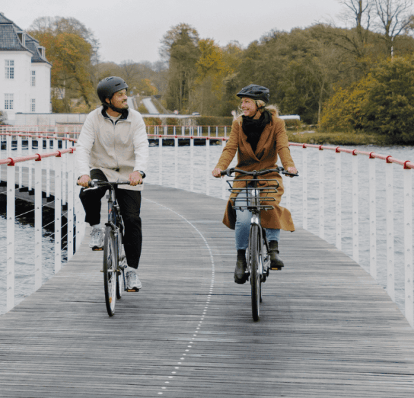 Two cyclists are riding on a bike path built on water, with a white manor house in the background