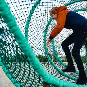 Et større barn inde i 'fiskeruse' på en legeplads med faldsand. I baggrunden ses et legetårn.