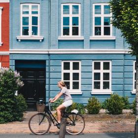 Kvindelig cyklist foran en række af byhuse; et rødt, et blåt og et gråt.