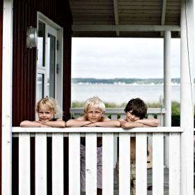 Tre børn titter op over et hvidt stakit i et sommerhus med udsigt ud til havet.