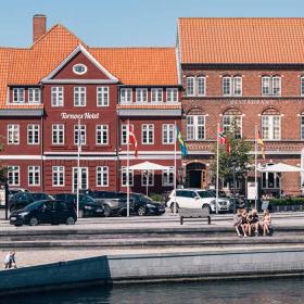 Store, røde huse foran havnefronten i Kerteminde. I forgrunden sidder der folk og nyder solen, en masse parkerede biler i baggrunden.