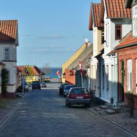 Brostensbelagt gade med gamle byhuse i forskellige farver til begge sider. Der er flag sat op foran et par af husene, et par parkerede biler, og den blå himmel ses i forsvindingspunktet.