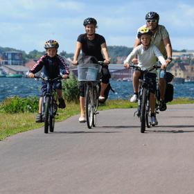 En familie på fire cykler sammen langs en sti ved kysten. Børnene fører an, og de voksne følger efter. I baggrunden ses det blå vand med en by helt i baggrunden. Det er en klar dag med solskin.