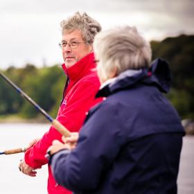 To lystfiskere står med fiskestængerne ved vandet. Den ene med ryggen til i en mørkeblå jakke, den anden kigger sig skråt over skulderen og er i en rød jakke. I baggrunden ses vandet og grøn beplantning inde på land.