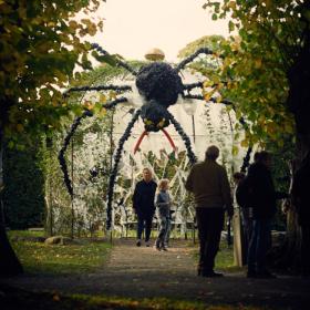 Mellem to træer står en kvinde og en pige under en kuppelformet pavillon. På taget kravler en stor edderkop lavet af planter og blade.  