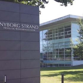 På et højt skilt står der Nyborg Strand Hotel og Konference. Bagved skiltet kan man se en græsplæne med tre træer der står foran en hvid firkantet bygning med mange vinduer.