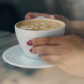 Gennem et vindue er der taget et nærbillede af to hænder der holder en kop kaffe. Hænderne har lyserød neglelak på. Hænderne holder om en hvid kop. I koppen er der kaffe og den er pyntet med latte-art der ligner et blad. Koppen står på en hvid underkop, som står på et lyst træbord.