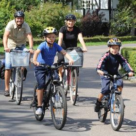 To voksne og to børn cykler af sted på stille asfaltvej med bindingsværkshus i baggrunden. Øverst i højre hjørne ses det røde Bike Friend-logo.