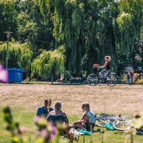 Munkemose park i Odense på en sommerdag. En flok unge mennesker sidder på et tæppe og hygger sig, mens en cyklist cykler på en sti i baggrunden.