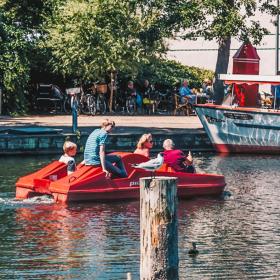 Odense Aafart Bådudlejning på en solskinsdag. En familie kommer sejlende i en vandcykel og mange mennesker sidder ude foran Nelle's i det gode vejr og kigger ud på åen..  