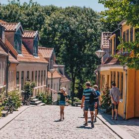 Gammel stræde med brosten og bindingsværkshuse i orange, røde og beige farver. En flok mennesker går tur i det gode vejr og kigger på husene.