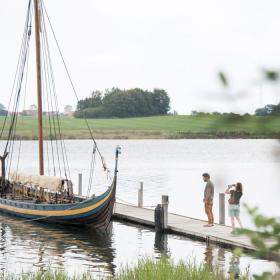 Det meste af billedet er består af vand som i horisonten mødes med græs. Midt i billedet ses en badebro, hvor der ligger et gammelt vikingeskib bundet fast. På badebroen står en kvinde og en mand. Kvinden fotograferer skibet.