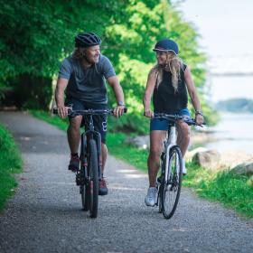 I venstre side cykler et par på hver deres cykel og cykelhjelm. De cykler på en sti der fører ud af en skov. Til højre i billedet adskiller en række af sten havet fra stien. I horisonten kan man se den gamle Lillebæltsbro. 