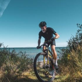 Person på cykel på vej op ad stejl skrænt med havet i baggrunden.