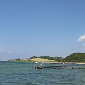 Kig over vand med badebro og strand og marker i baggrunden
