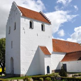 Foran en blå himmel står en hvis kirke med rødt tag. Den er omgivet af en kirkegård.