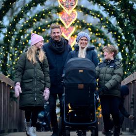 På en træbro står en familie med tre brøn i vintertøj. Foran moren står der en barnevogn. Over hele broen er der buer med gran, lyskæder og i midten et lysende julehjerte.