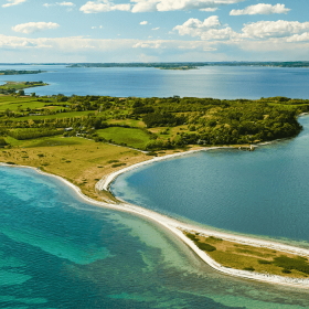 Dronefoto af Avernakø i skarpe, grønne farver med dybblåt vand hele vejen rundt om og horisonten i baggrunden. 