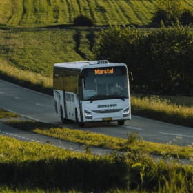 En hvid bus kører blandt grønne marker ned ad bakke på en sommerdag