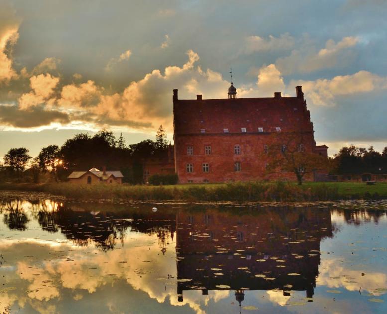 Spejling af det smukke røde slotshotel Broholm Slot i sø med oplyste skyer som baggrund.