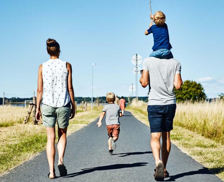 En familie på fire går ned ad en landevej på en solrig dag. Manden til højre bærer et lille barn på sine skuldre. Mellem dem går et andet barn. De er omgivet af åbne marker, og himlen er klar og blå.