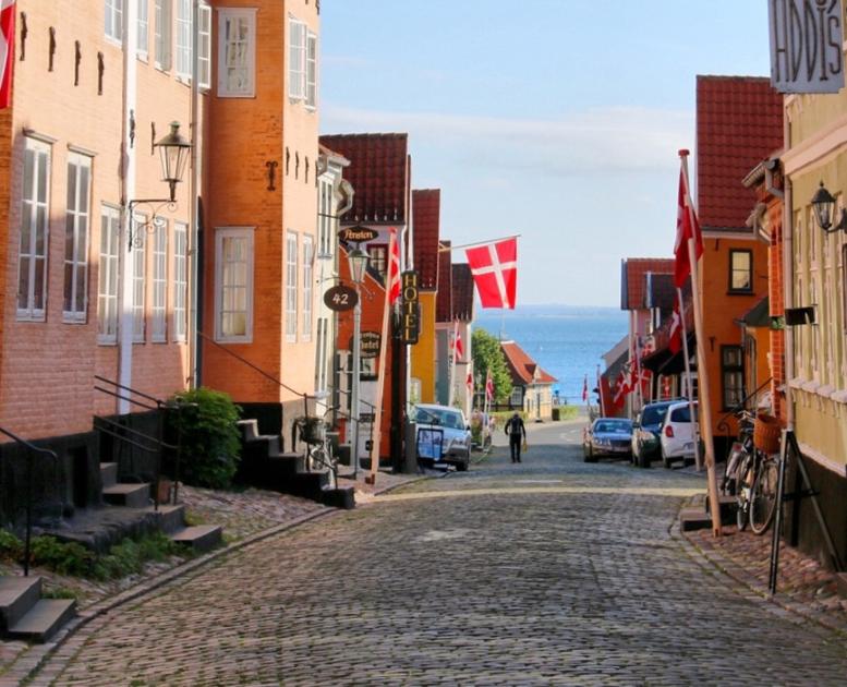 Brostentbelagtgade i Ærøskøbing om sommeren med havkig. Stokroser i forgrunden og farverige huse.