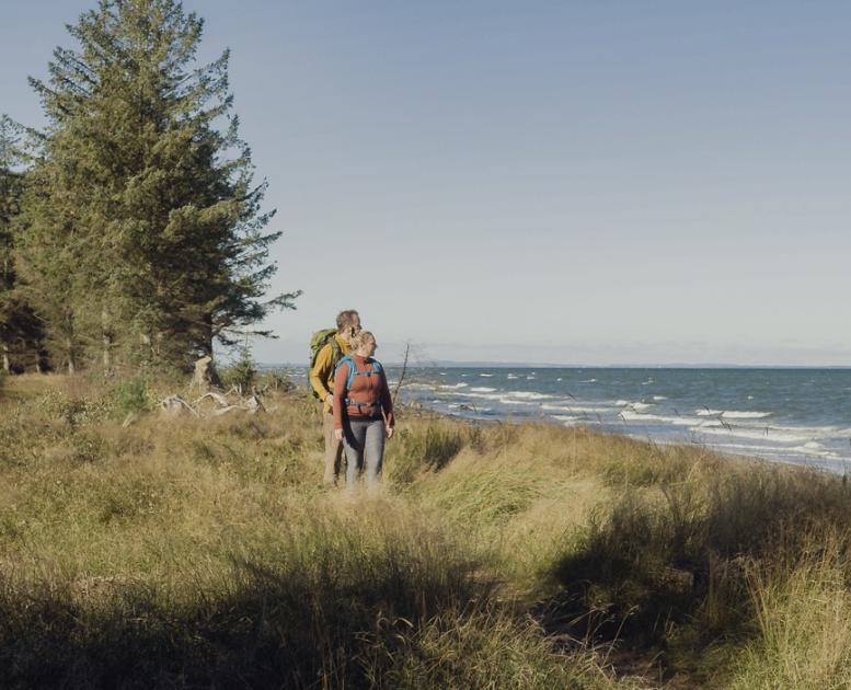 Kvinde og mand vandrer på græsklint ved kysten med grantræer i baggrunden.