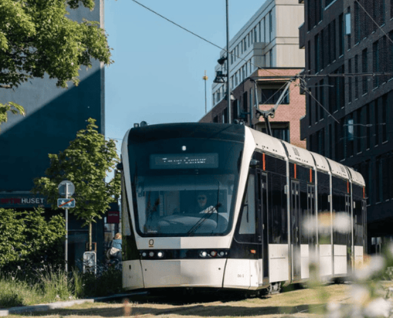 En letbane kører igennem en by med grønne træer på en solrig dag i Odense
