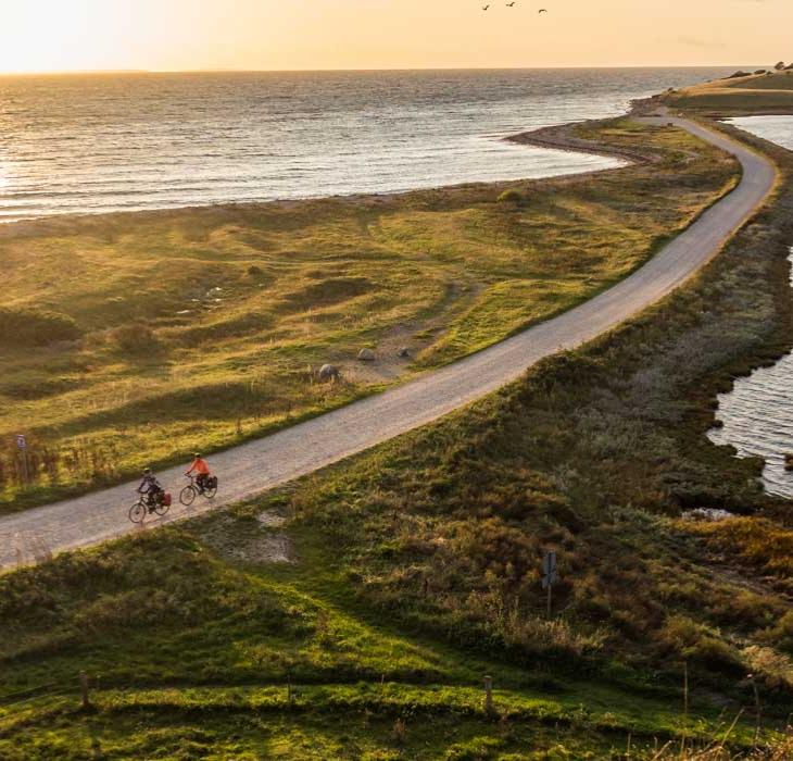 To cyklister cykler på smal grussti lige ud til vandet ved en fynsk strand. I baggrunden ses en smuk solnedgang. 