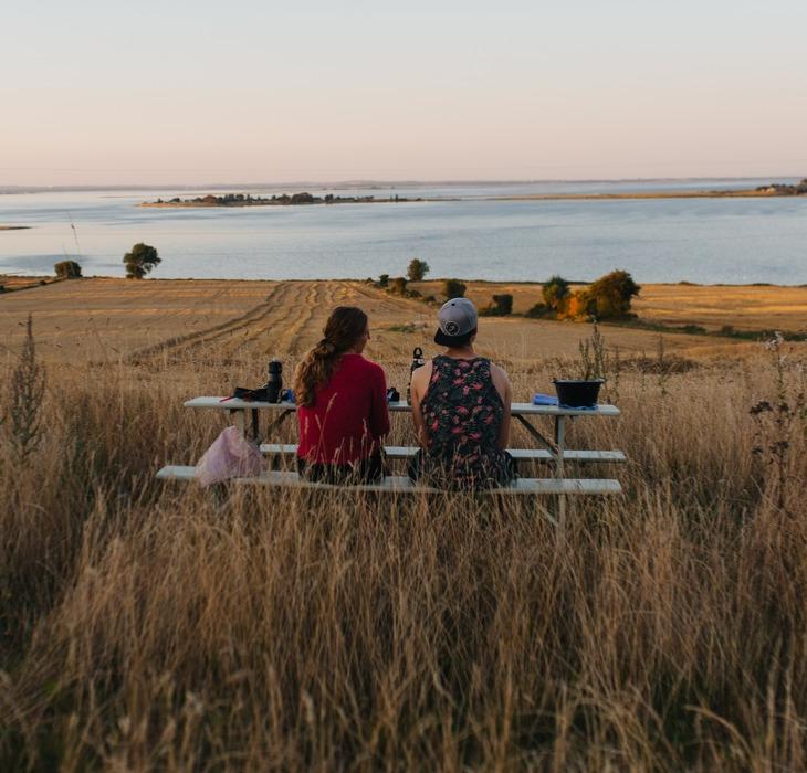 På en mark med udsigt udover havet står et bordebænkesæt. På bænken sidder to kvinder og kigger udover vandet.