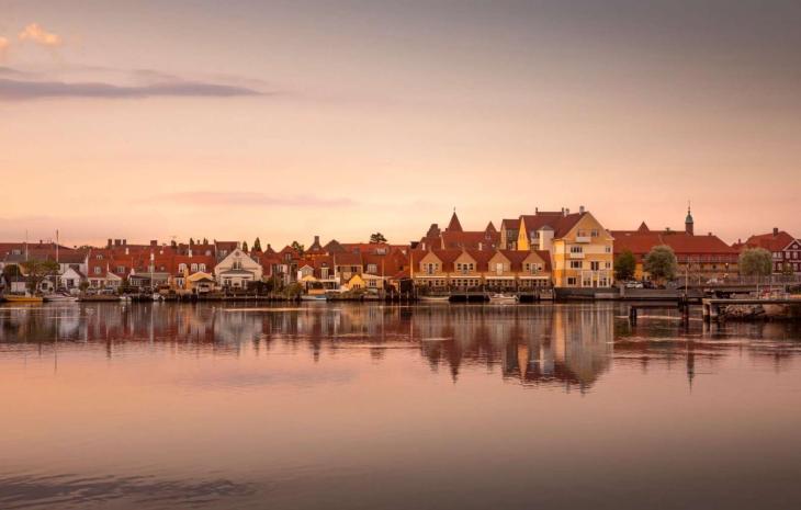 Kerteminde by set fra vandet. Solen er ved at gå nå så himlen er delvist lyserød. I forgrunden er der vand og i bagrunden ses Kerteminde by. Den består af en masse små bygninger i forskellige farver. De har dog alle røde tage.
