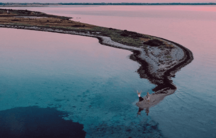 Dronefoto af en aflang landtange, hvor der står to mennesker på, med solnedgangsfarver i baggrunden