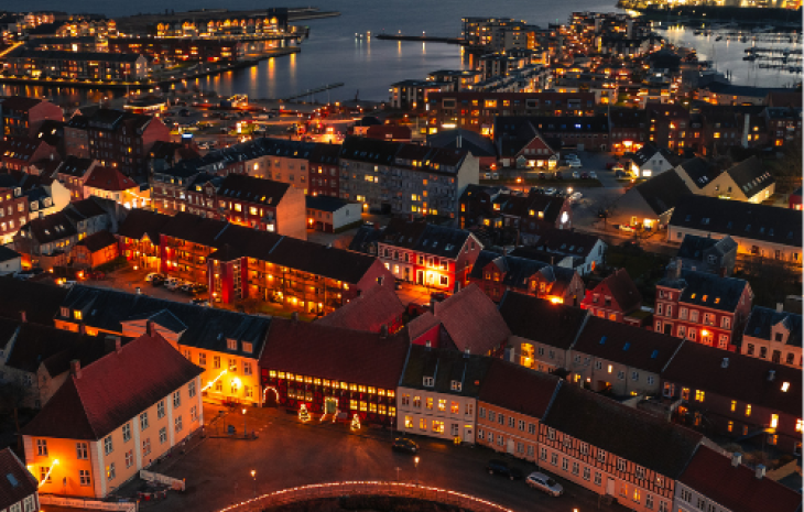 Dronefoto af Nyborg med sø og hav på hver sin side, gadebelysning og et skumringslys i horisonten